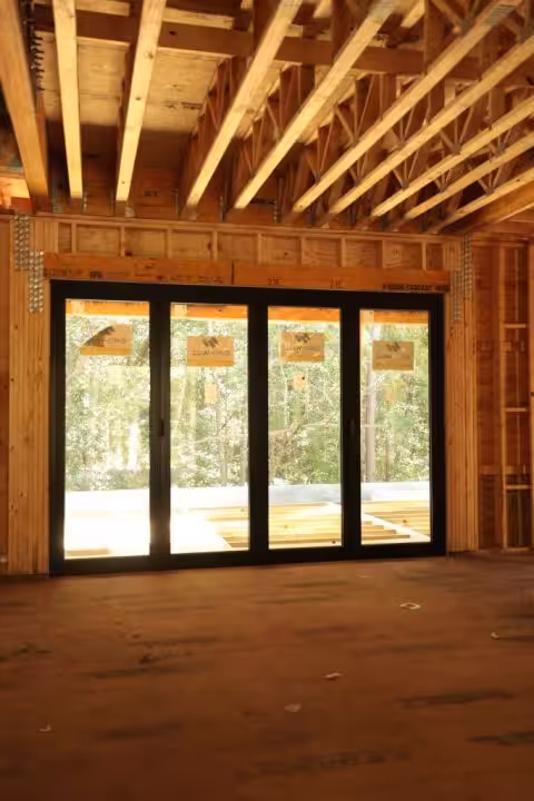 Inside a framed house, a deep header spans a four-panel glass door opening, with open-web floor trusses running overhead